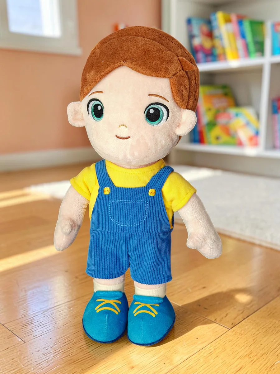Levi, a Playnitive fiú baba egy gyerekszoba fapadlóján állva: barna haj, sárga póló és kék kordbársony kertésznadrág